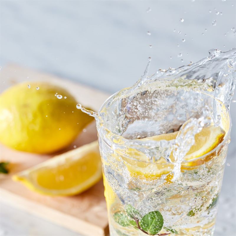 Water splashing in a glass of lemon and mint, beside a whole lemon and a lemon wedge on a wooden board.