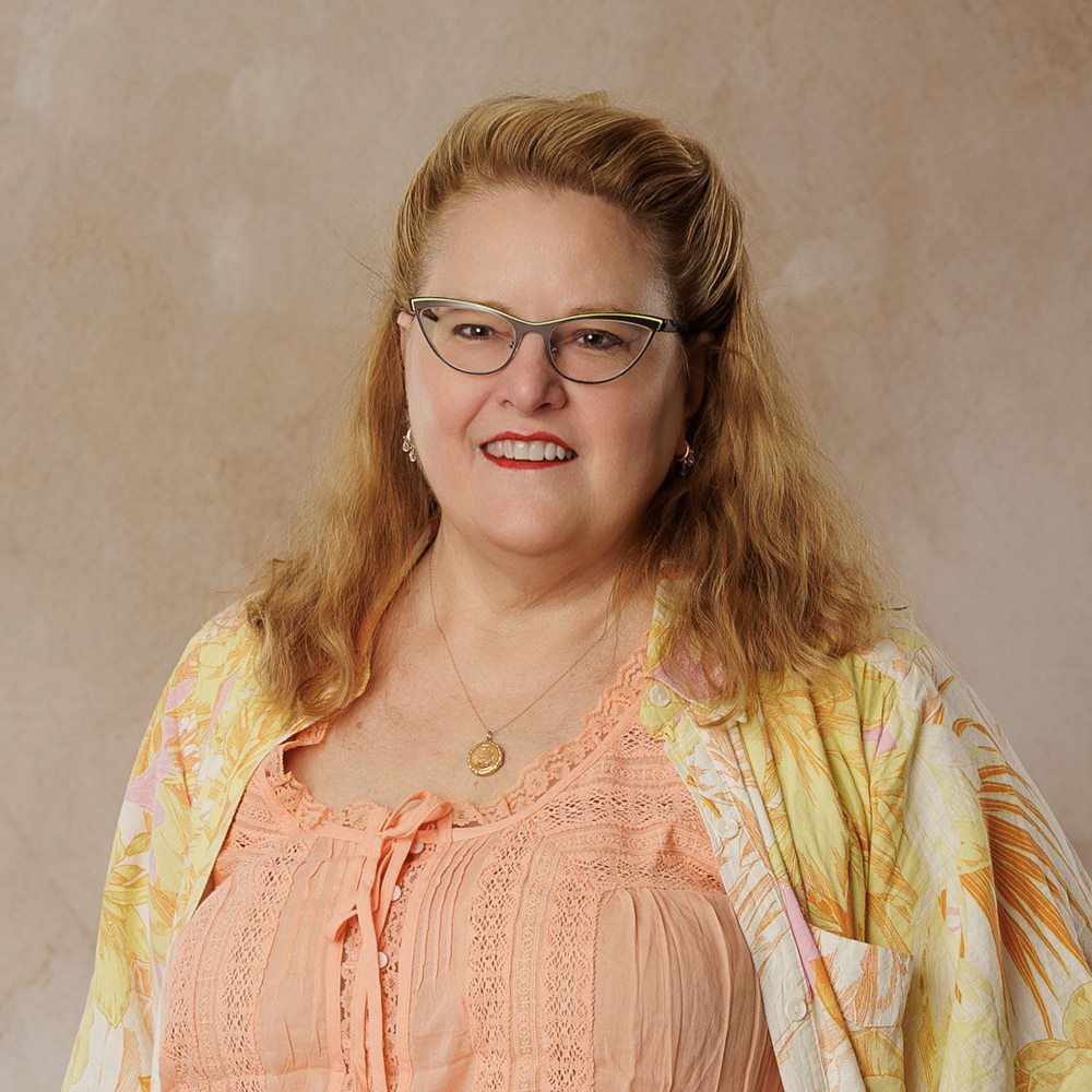 Sally Seaver, smiling, wearing glasses and a patterned robe over a pale top.