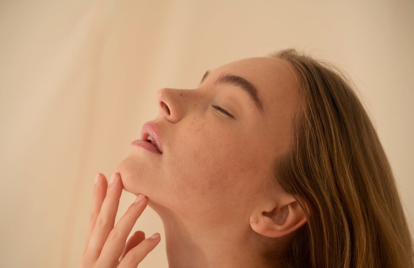 A woman with her eyes closed, her chin lifted and her fingertips resting beneath her jaw, in warm daylight.